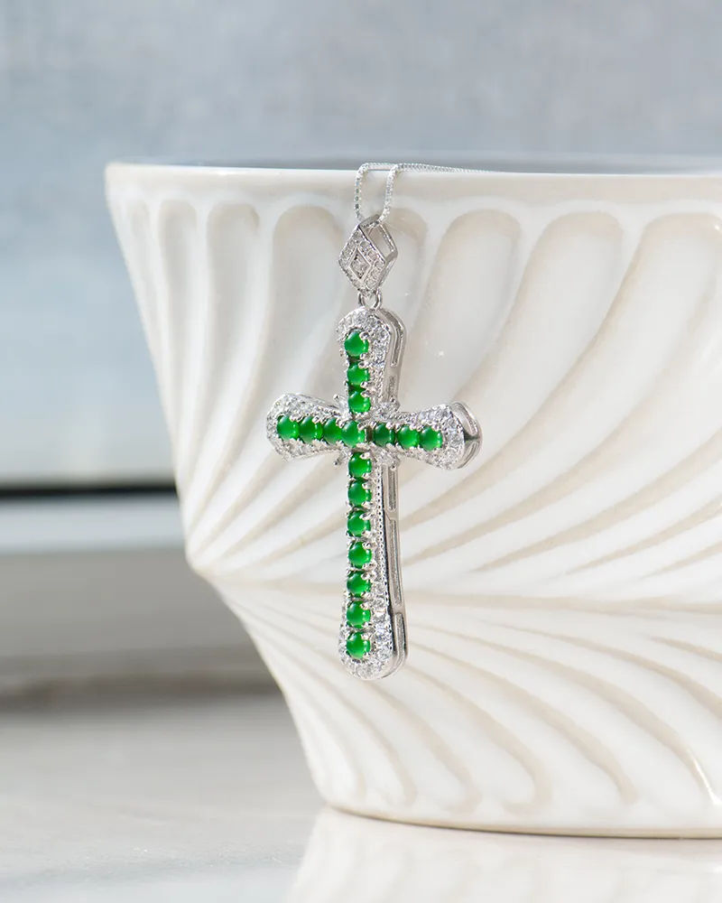 A silver cross pendant with green gemstones and small clear stones hangs against a white, textured ceramic bowl. The background is softly blurred, highlighting the jewelry.