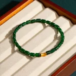 A green beaded bracelet with a single gold bead is displayed on a cream-colored, cushioned jewelry tray in a wooden box.