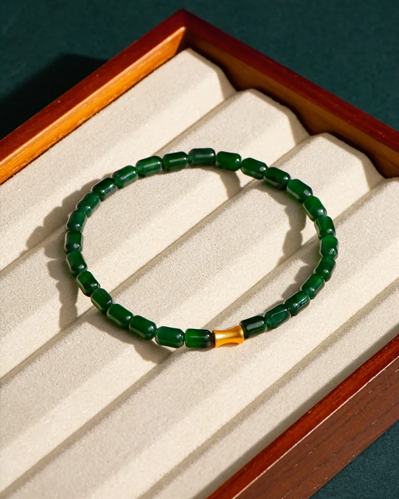 A green beaded bracelet with a single gold bead is displayed on a cream-colored, cushioned jewelry tray in a wooden box.