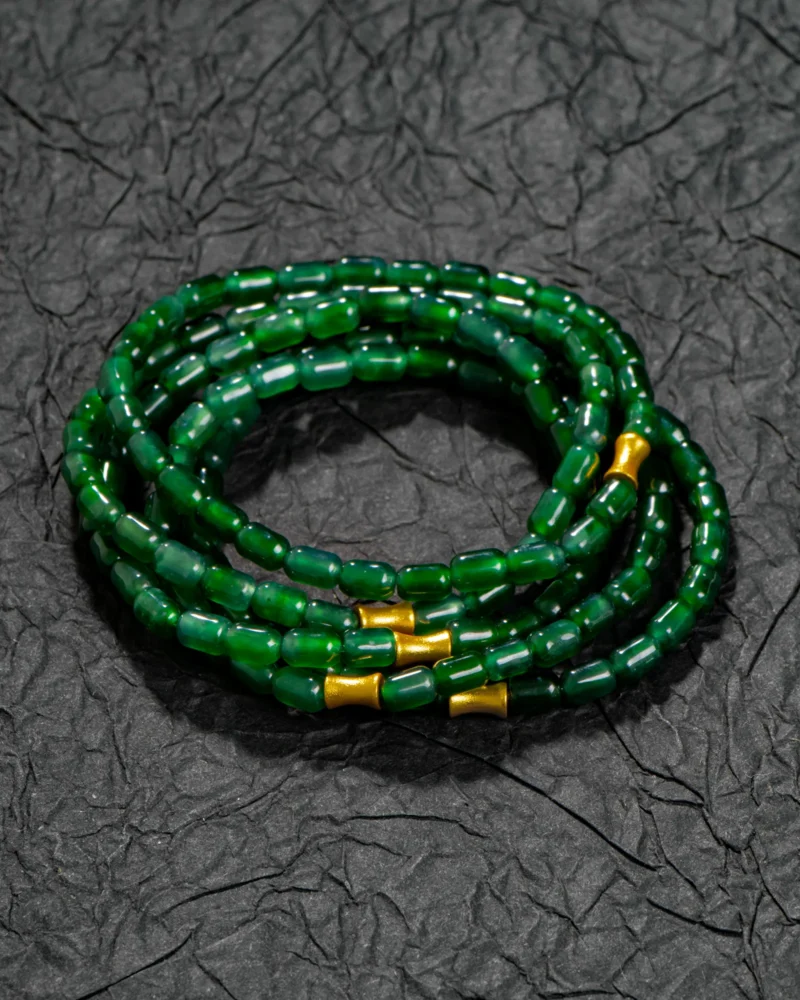 The Deep Green Feicui Bracelet features glossy, drum-shaped green beads accented with three warm-tone spacers, artfully displayed on a textured black surface.