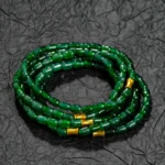A coil of deep green glass beads with small gold accents, arranged in a stacked bracelet, rests on a textured black surface.