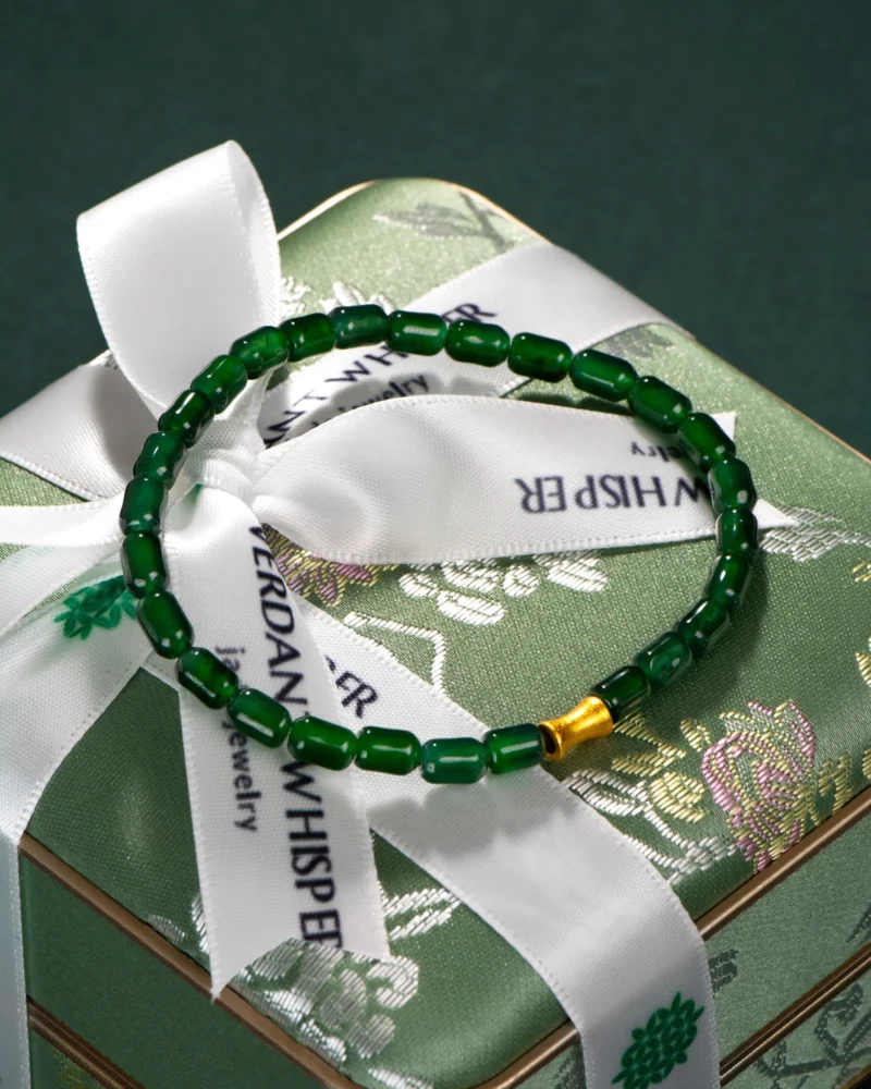 A Deep Green Feicui Bracelet featuring drum-shaped beads and a warm-tone spacer rests atop a green floral-patterned gift box tied with a white ribbon.