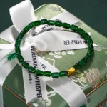 A green beaded bracelet with a single gold bead is displayed on a green floral-patterned gift box wrapped with a white ribbon printed with VERDANT WHISPER Jewelry in black text.