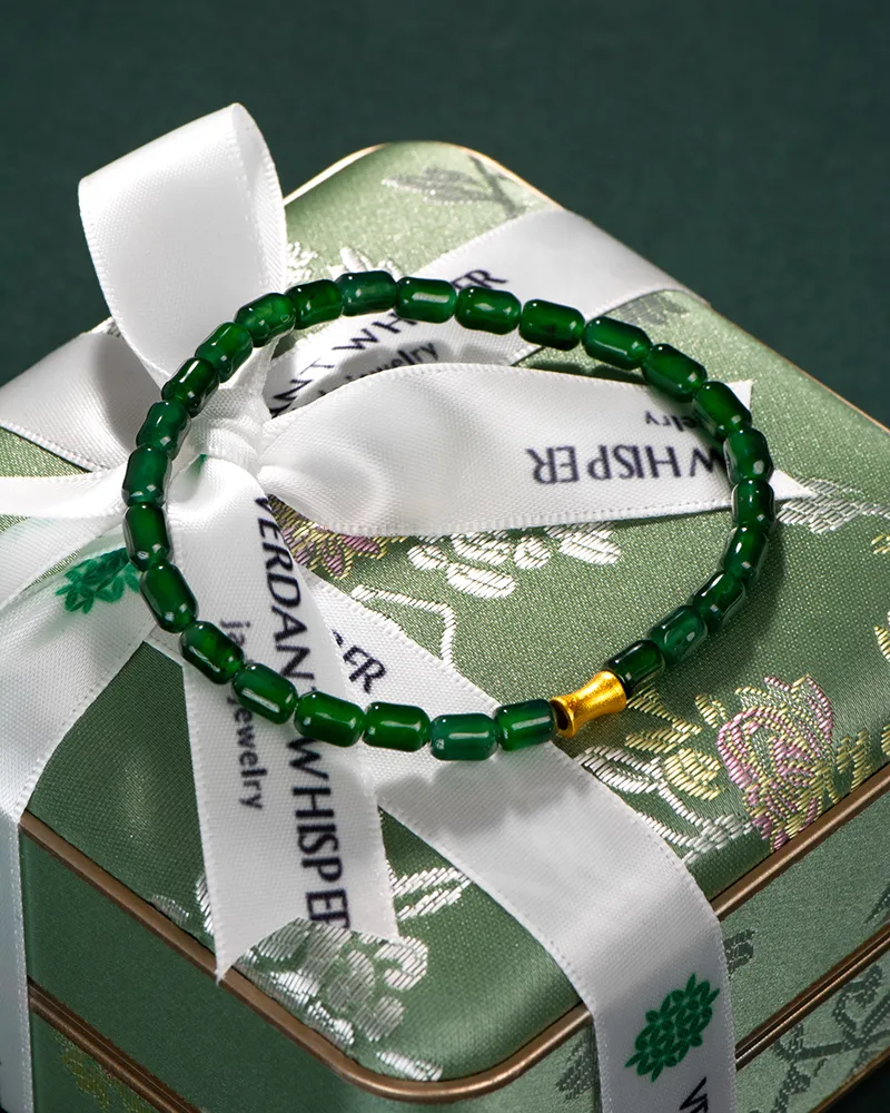 A green beaded bracelet with a single gold bead is displayed on a green floral-patterned gift box wrapped with a white ribbon printed with VERDANT WHISPER Jewelry in black text.