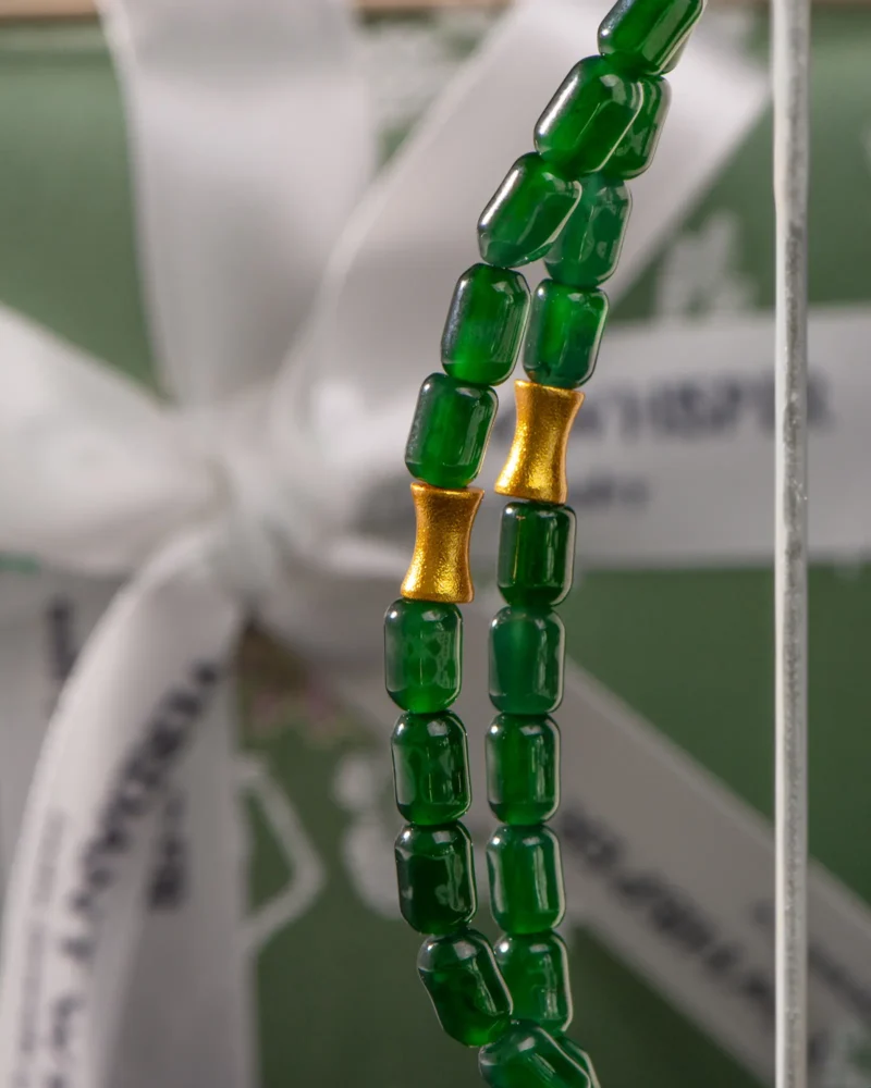Close-up of the Deep Green Feicui Bracelet, showcasing its cylindrical drum-shaped beads and two warm-tone gold spacers, elegantly displayed in front of a softly blurred green gift box tied with a white ribbon.