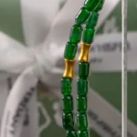 Close-up of green glass beads and two gold cylindrical beads, strung together, with a blurred white ribbon and green background behind them.