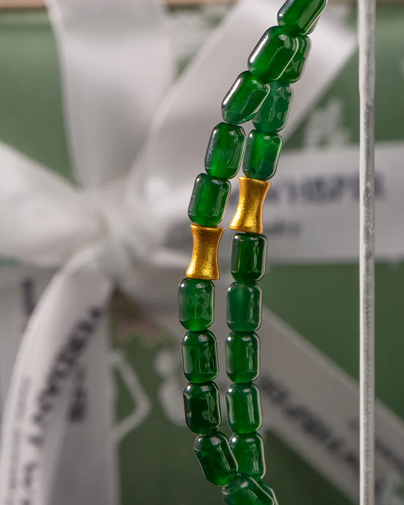Close-up of green glass beads and two gold cylindrical beads, strung together, with a blurred white ribbon and green background behind them.