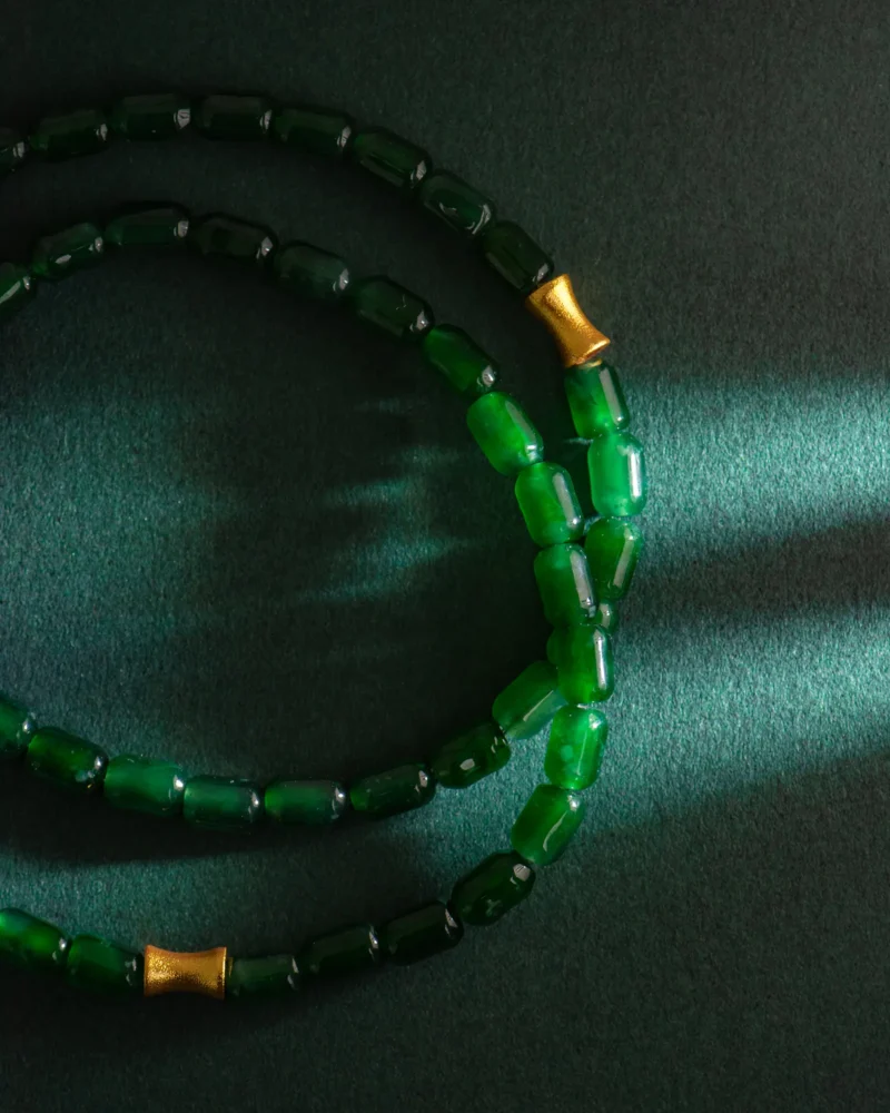 Two strands of the Deep Green Feicui Bracelet—featuring drum-shaped beads in a rich jade hue and accented with warm-tone cylindrical spacers—are arranged in circles on a textured dark green surface, partially illuminated by a shaft of light.