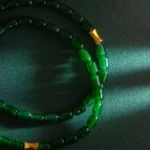 Two strands of green jade beads with small gold accents lie on a dark green surface, illuminated by a slant of light from the right side, casting soft shadows.