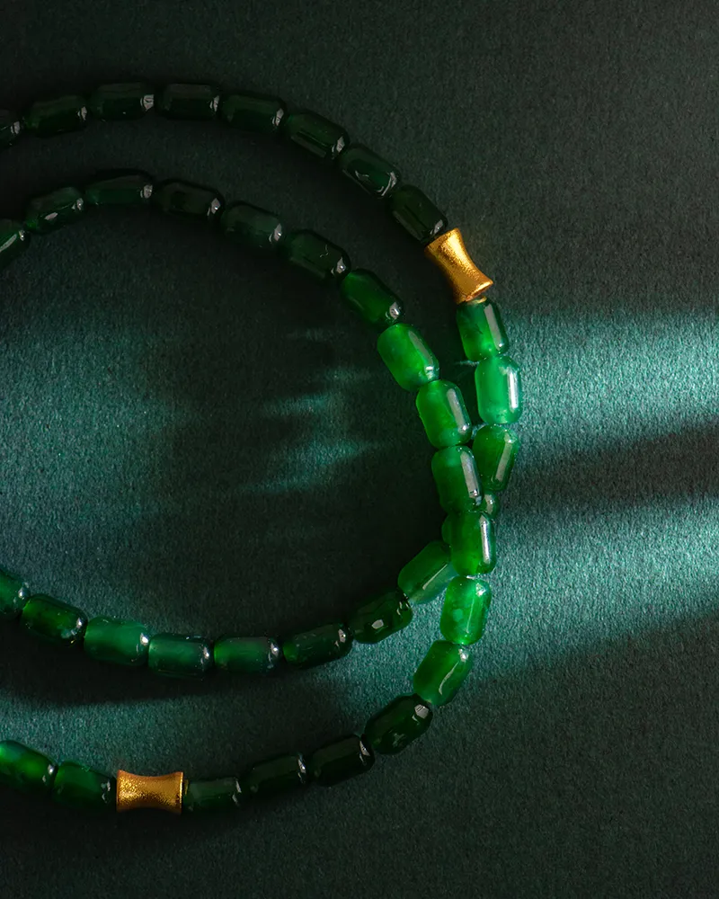 Two strands of green jade beads with small gold accents lie on a dark green surface, illuminated by a slant of light from the right side, casting soft shadows.