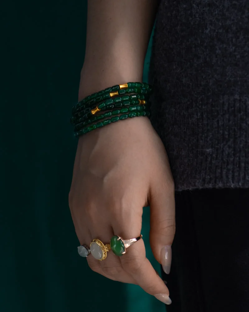 A hand with long nails wears three gemstone rings and the Deep Green Feicui Bracelet, which features drum-shaped beads and warm-tone spacers arranged in multiple strands. The individual is dressed in a dark gray sweater and black pants against a dark green background.