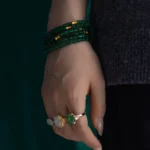 A hand wearing three rings with green and white stones, and a multi-strand dark green beaded bracelet. The person is dressed in a dark top and black pants.