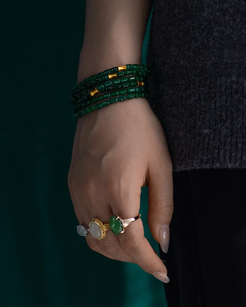 A hand wearing three rings with green and white stones, and a multi-strand dark green beaded bracelet. The person is dressed in a dark top and black pants.