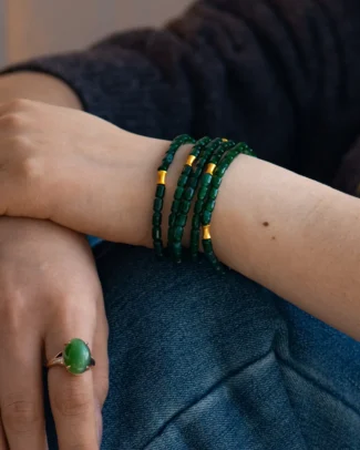 A close-up of a person’s wrist adorned with the Deep Green Feicui Bracelet, featuring drum-shaped beads and warm-tone spacers. The person also wears a silver ring set with a large green stone and is dressed in dark clothing.