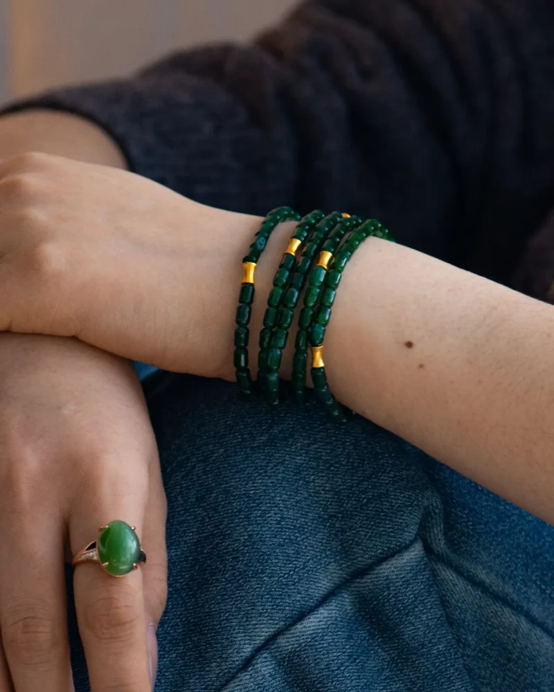 A close-up of a person’s wrist adorned with the Deep Green Feicui Bracelet, featuring drum-shaped beads and warm-tone spacers. The person also wears a silver ring set with a large green stone and is dressed in dark clothing.