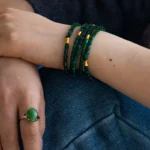 A person rests their hands on their knee, wearing multiple green bead bracelets with gold accents on one wrist and a large green gemstone ring on one finger. They are dressed in dark clothing and blue denim.