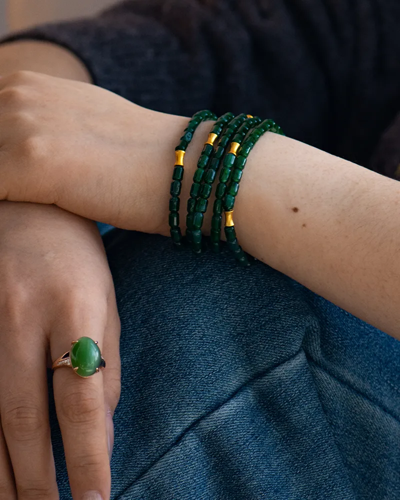A person rests their hands on their knee, wearing multiple green bead bracelets with gold accents on one wrist and a large green gemstone ring on one finger. They are dressed in dark clothing and blue denim.