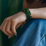 A person wearing blue jeans and a brown top rests their hand on their knee, showing multiple green and yellow beaded bracelets wrapped around their wrist.