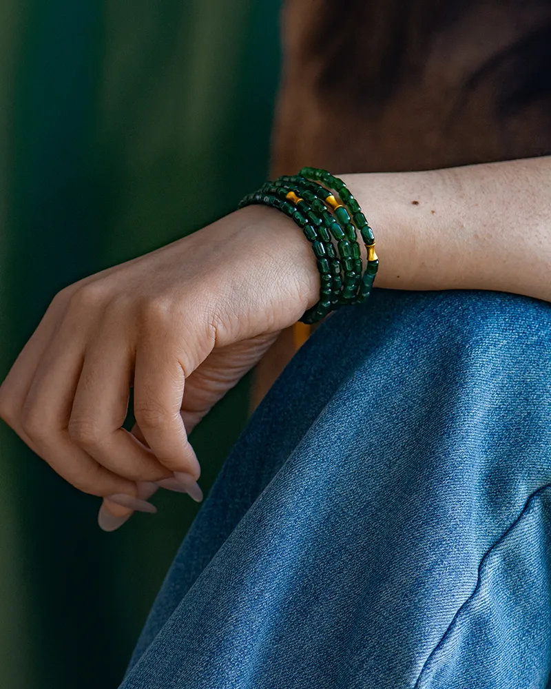 A person wearing blue jeans and a brown top rests their hand on their knee, showing multiple green and yellow beaded bracelets wrapped around their wrist.