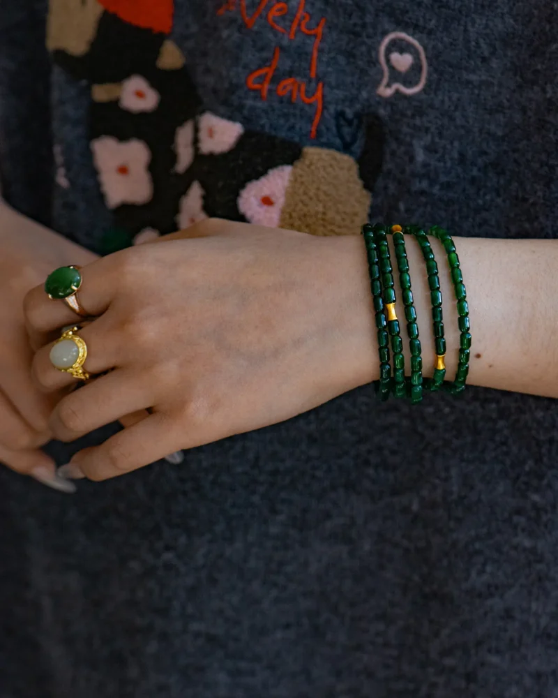A hand wearing two rings and the Deep Green Feicui Bracelet — featuring drum-shaped beads with a warm-tone spacer — rests against dark fabric adorned with a colorful floral pattern and the partially visible words “every day.”.