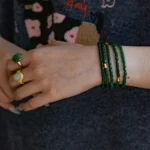 A person’s hand wearing two gemstone rings and several green beaded bracelets rests against a dark shirt with a floral and red text pattern.