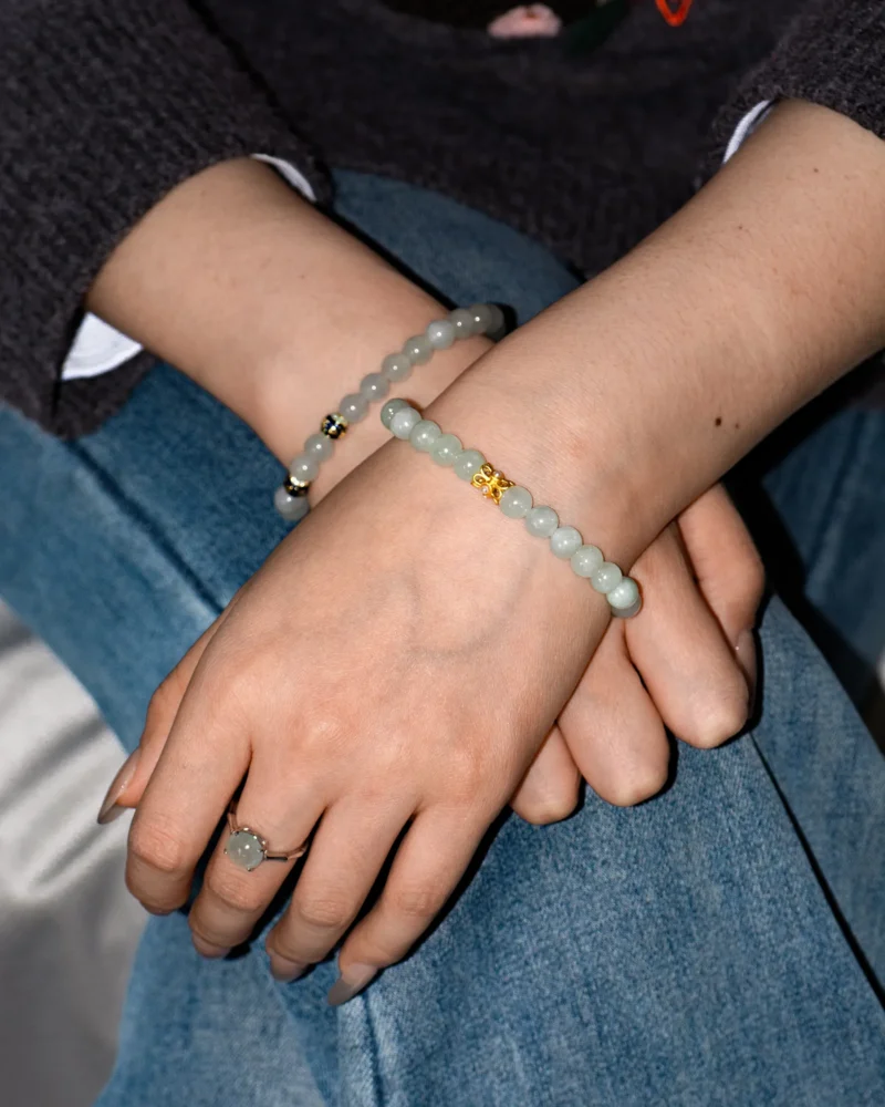 A person wearing blue jeans and a dark sweater crosses their hands, highlighting the Light Green Jadeite Bead Bracelet — featuring spherical jadeite beads with warm accents — beautifully paired with a matching jade ring on their finger.