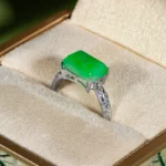 A silver ring with a rectangular green gemstone, possibly jade, set in the center, displayed in a beige, cushioned jewelry box with a fabric-covered exterior.