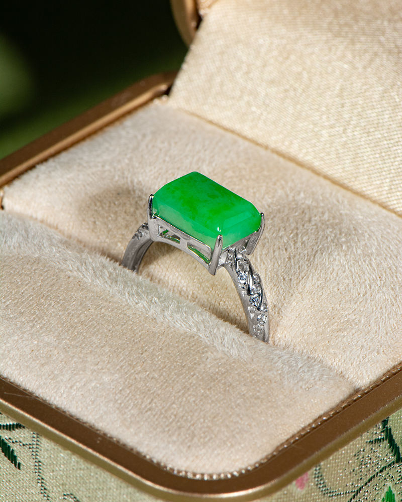 A silver ring with a rectangular green gemstone, possibly jade, set in the center, displayed in a beige, cushioned jewelry box with a fabric-covered exterior.