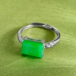 A silver ring featuring a rectangular green jade stone as the centerpiece, accented with small clear stones on the band, is displayed on a light green textured surface.