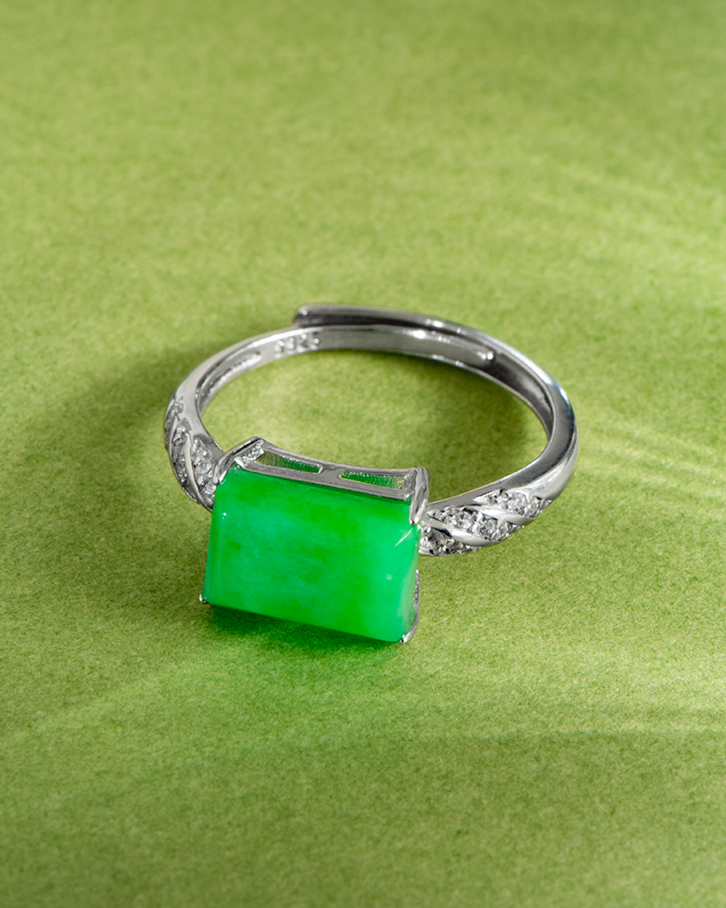 A silver ring featuring a rectangular green jade stone as the centerpiece, accented with small clear stones on the band, is displayed on a light green textured surface.