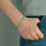 A person wearing a grey top and blue jeans has a hand resting on their hip, showcasing a silver bracelet with five large green gemstones, and a simple gold ring on their finger.