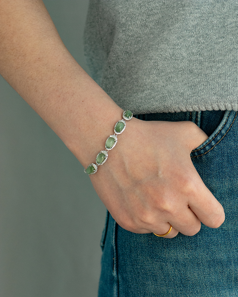 A person wearing a grey top and blue jeans has a hand resting on their hip, showcasing a silver bracelet with five large green gemstones, and a simple gold ring on their finger.