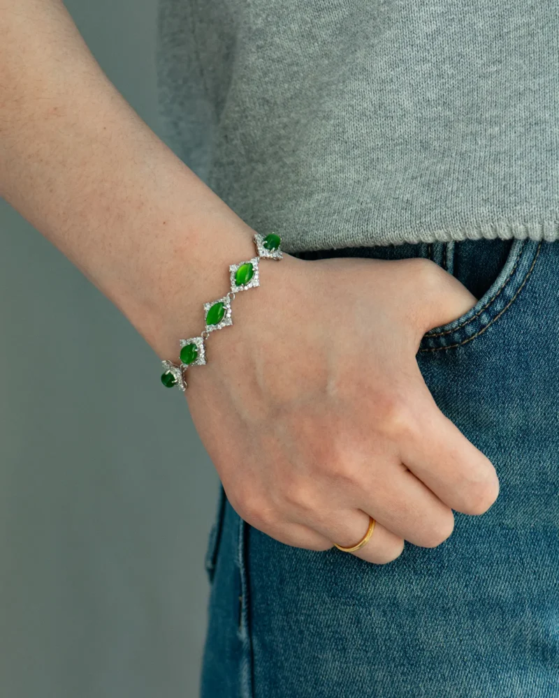 A person wearing a gray top and blue jeans is accessorizing with a gold ring on their finger and the Green Nephrite Jade Bracelet — Marquise Cabochon Link Line on their wrist.