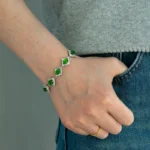 A person wearing a gray top and blue jeans has their hand in their pocket, showing a gold ring and a silver bracelet with green gemstones and surrounding white stones on their wrist.