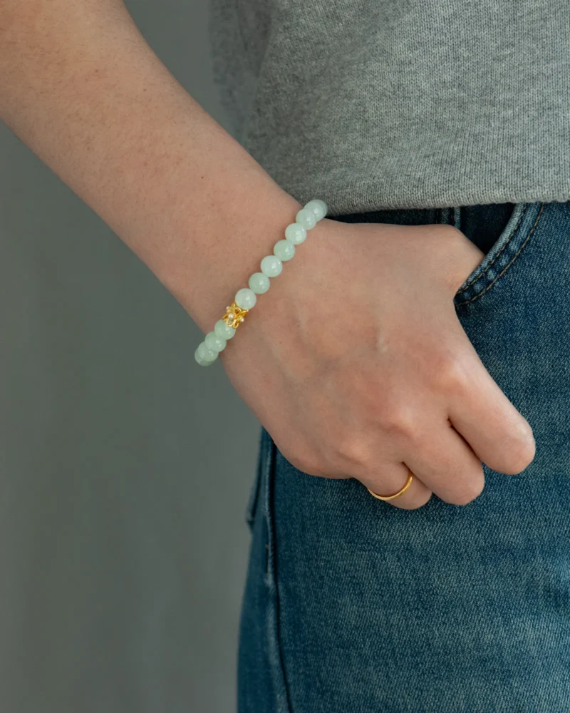 A person wearing the Light Green Jadeite Bead Bracelet — featuring spherical beads with a gold-tone detail — and a gold ring, with one hand in the pocket of blue jeans, set against a gray background.