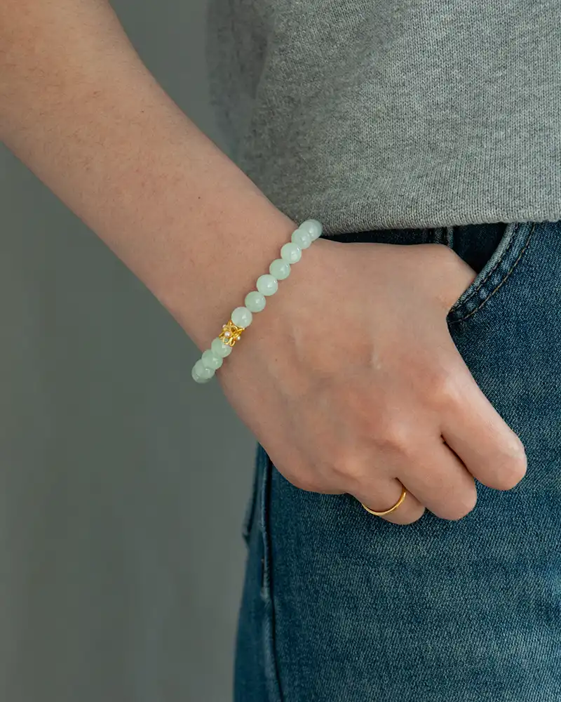 LYU_5657 A person wearing a light green beaded bracelet with a gold accent, a gold ring, a gray top, and blue jeans. The hand rests casually by their side.