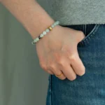 A person wearing a gray shirt and blue jeans has their left hand in their pocket, displaying a gold ring and a beaded bracelet with white and decorative beads on their wrist.