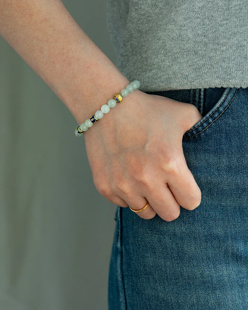 A person wearing a gray shirt and blue jeans has their left hand in their pocket, displaying a gold ring and a beaded bracelet with white and decorative beads on their wrist.