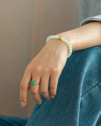 A hand wearing the Light Green Jadeite Bead Bracelet — featuring spherical beads with a gold-tone detail — along with a silver ring set with a green stone, rests on a knee clad in blue jeans.