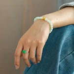 A person wearing a light jade beaded bracelet with a gold accent and a green rectangular stone ring rests their hand on their blue-jeaned knee.