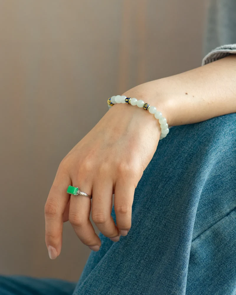 A hand wearing a green stone ring and the Light Green Jadeite Bead Bracelet—spherical beads with warm accents—rests on denim-clad legs.