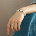A person wearing blue jeans rests their hand on their leg, displaying a silver ring with a green stone and a beaded bracelet with white and gold accents on their wrist.