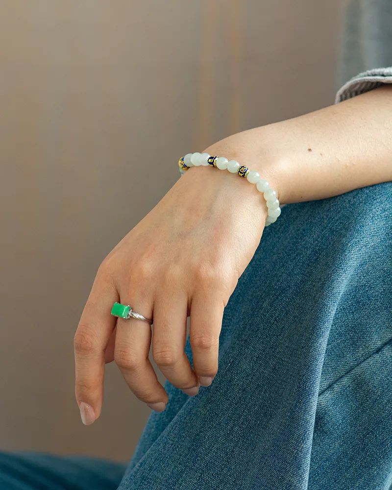 A person wearing blue jeans rests their hand on their leg, displaying a silver ring with a green stone and a beaded bracelet with white and gold accents on their wrist.