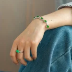 A person wearing blue jeans rests their hand on their knee, showing a silver bracelet with green stones and a matching green stone ring on their finger.