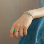 A person wearing blue jeans displays a silver bracelet with green stones on their wrist and a matching green stone ring on their finger. The hand rests gently on their leg.