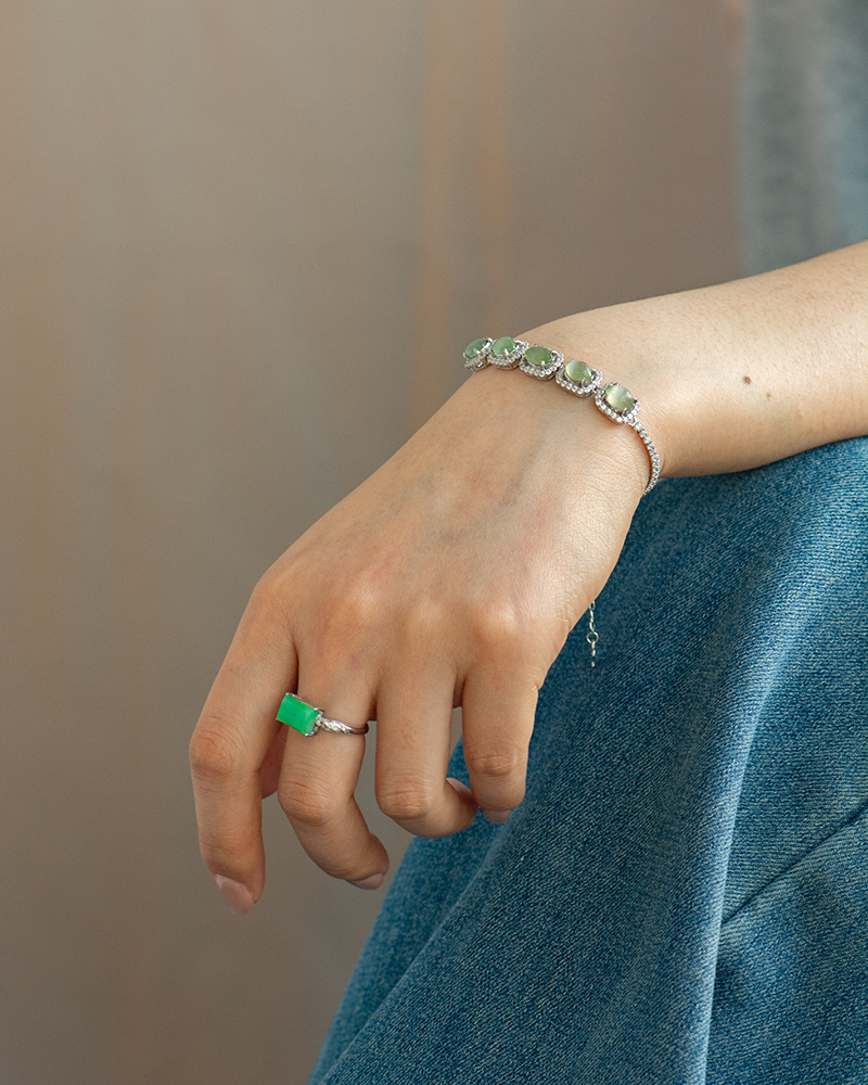 A person wearing blue jeans displays a silver bracelet with green stones on their wrist and a matching green stone ring on their finger. The hand rests gently on their leg.
