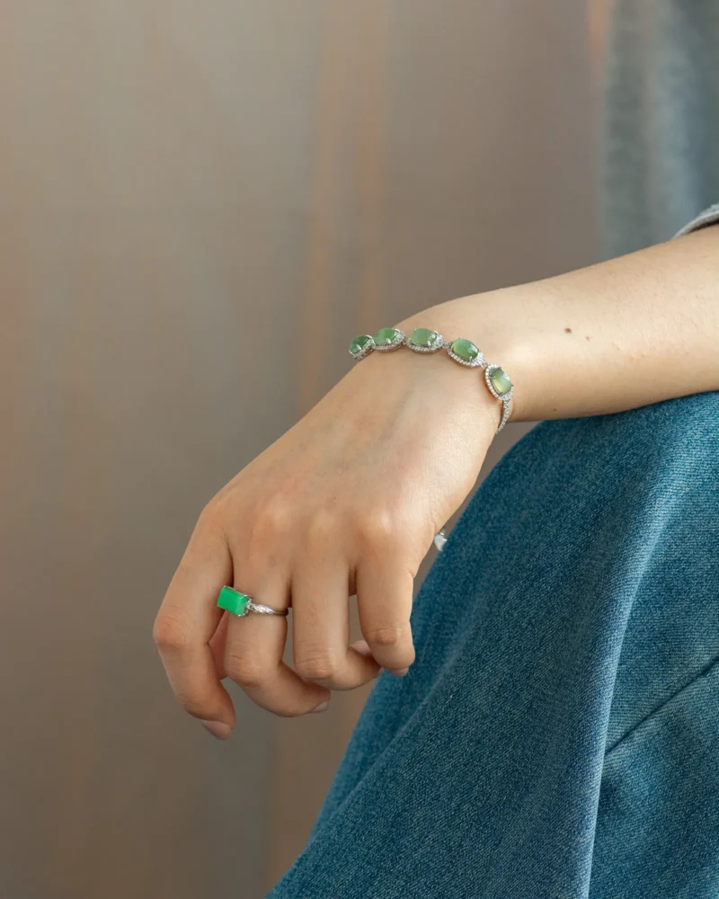 A hand resting on a knee is adorned with the Grayish-White Nephrite Jade Bracelet — Oval Cabochon Links, featuring oval green gemstones, paired with a matching silver ring set with a large green stone. The person is dressed in blue jeans and a grey top.