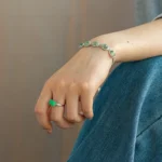 A close-up of a persons hand wearing a silver bracelet with green gemstones and a matching green gemstone ring, resting on their blue-jeaned lap.