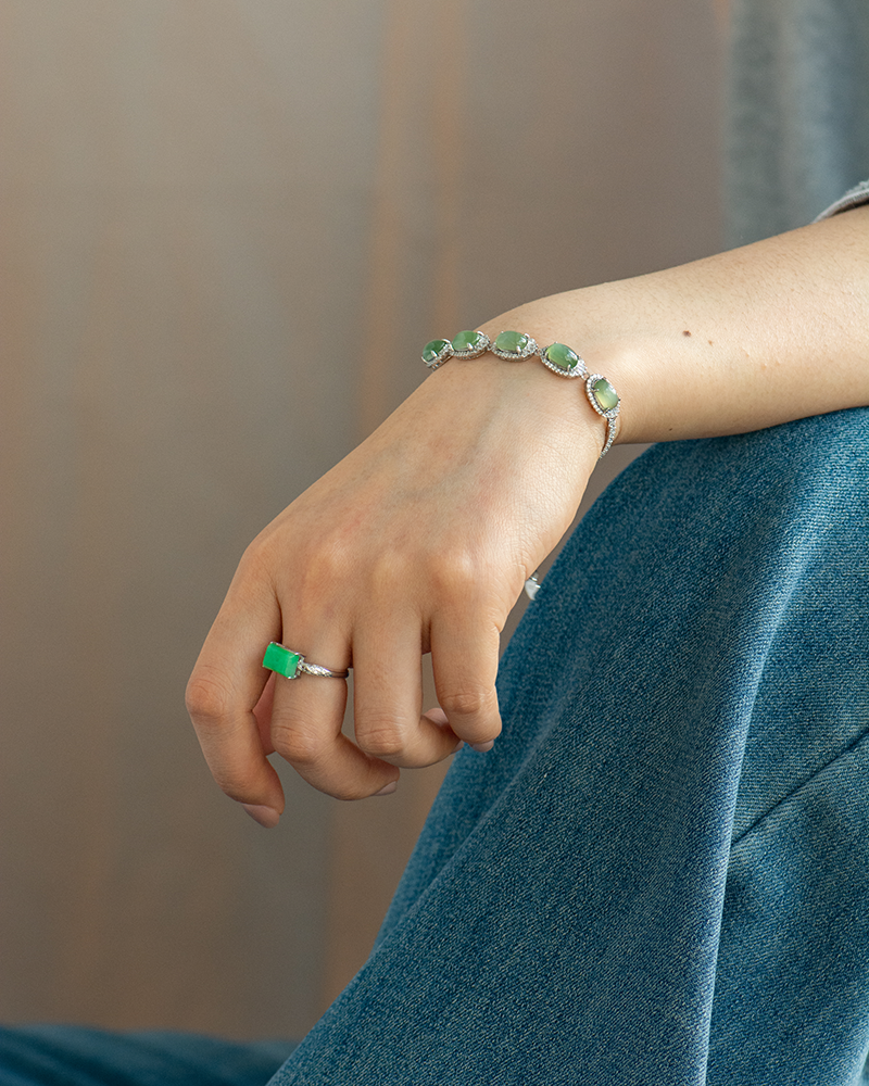 A close-up of a persons hand wearing a silver bracelet with green gemstones and a matching green gemstone ring, resting on their blue-jeaned lap.
