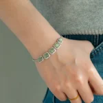 A person wearing a grey top and blue jeans rests their hand in their pocket. A silver bracelet with five green gemstone settings and a thin gold ring are on their wrist and finger.